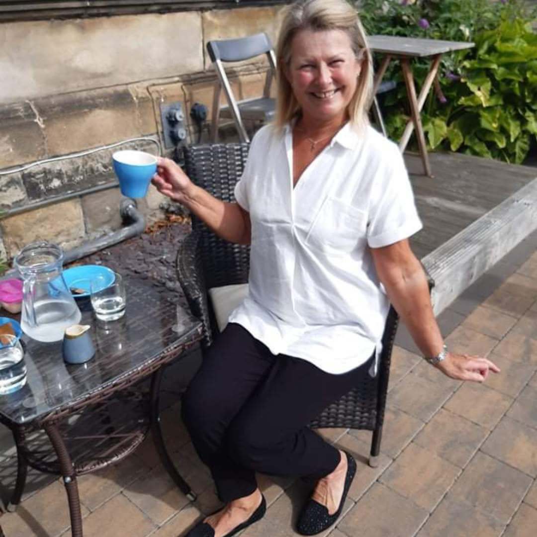 Woman sitting outdoors holding a blue cup, with a table and chairs in the background.